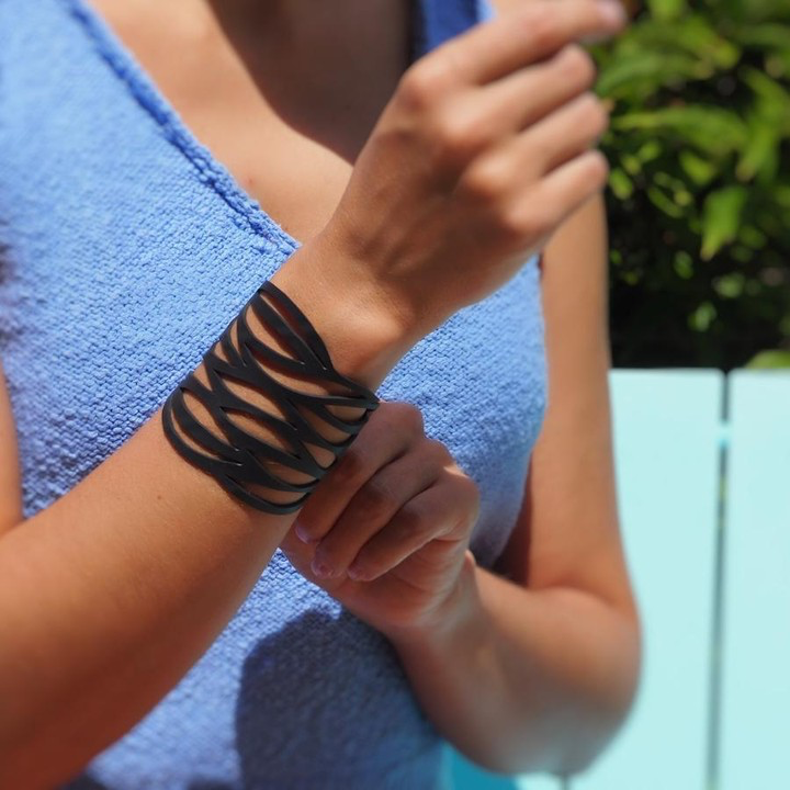 Paguro Upcycle black cuff recycled rubber bracelet with wave cutouts worn on a womans wrist with blue top outdoors